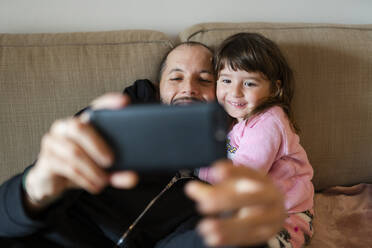 Happy father and daughter making a video call to the family with the mobile phone, sitting on the couch - GEMF03497