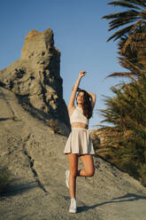 Porträt einer modischen jungen Frau, die auf einem Felsen steht und den Sonnenuntergang genießt - MPPF00657