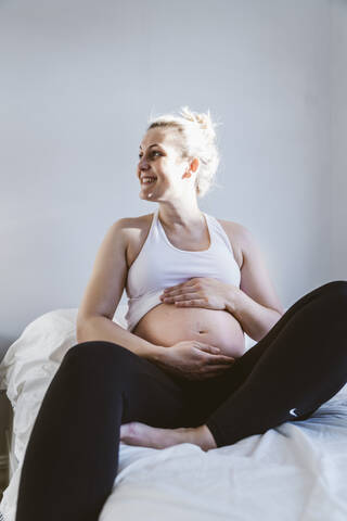 Glückliche schwangere Frau sitzt auf dem Bett zu Hause, lizenzfreies Stockfoto