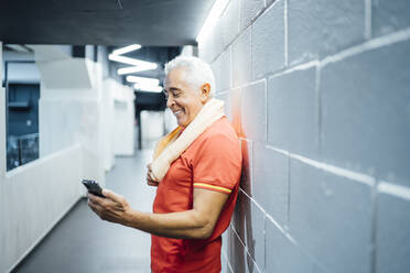 Lächelnder älterer Mann, der eine Pause macht und sein Handy im Fitnessstudio benutzt - OCMF01115