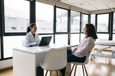 Lächelnder Arzt mit Laptop im Gespräch mit einem Patienten in einer Arztpraxis - ABZF03014