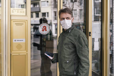 Mann mit Maske wartet vor einer Apotheke und hält ein Rezept in der Hand - MFF05131