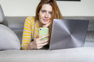 Portrait of young woman lying on the couch at home using smartphone and laptop - AFVF05771