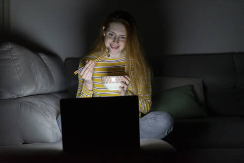 Junge Frau sitzt auf der Couch zu Hause und isst, während sie einen Laptop benutzt, lizenzfreies Stockfoto