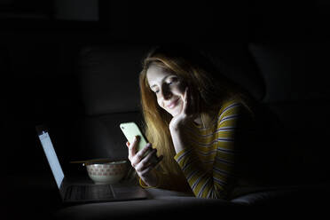 Young woman lying on the couch at home using laptop and smartphone - AFVF05783