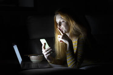 Junge Frau liegt zu Hause auf der Couch und benutzt Laptop und Smartphone - AFVF05784