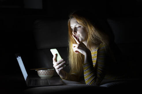 Young woman lying on the couch at home using laptop and smartphone - AFVF05784