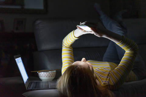 Junge Frau liegt zu Hause auf der Couch und benutzt ihr Smartphone, lizenzfreies Stockfoto