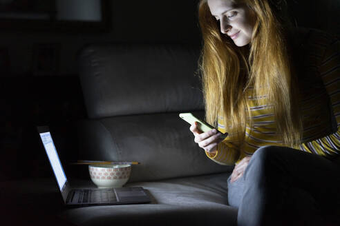 Young woman sitting on the couch at home using laptop and smartphone - AFVF05787