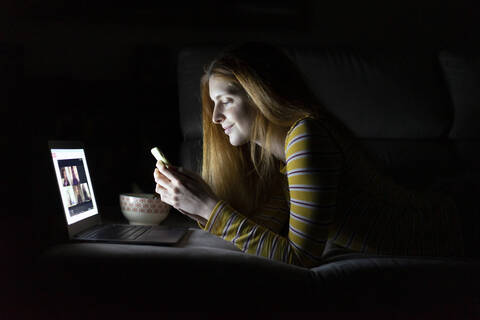 Junge Frau liegt zu Hause auf der Couch und benutzt Laptop und Smartphone, lizenzfreies Stockfoto