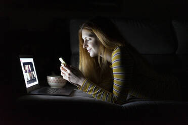 Junge Frau liegt zu Hause auf der Couch und benutzt Laptop und Smartphone - AFVF05790