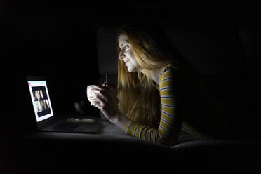 Young woman having a laptop video conference on the couch at home while having dinner - AFVF05792