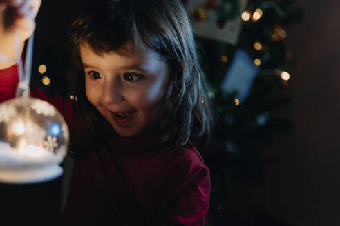 Porträt eines aufgeregten kleinen Mädchens, das eine beleuchtete Glaskugel zur Weihnachtszeit hält - GEMF03499