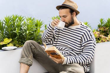 Mann sitzt auf dem Balkon, trinkt Kaffee und liest ein Buch - AFVF05798