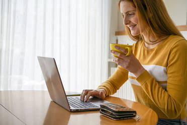 Lächelnde junge Frau mit einer Tasse Kaffee und einem Laptop zu Hause - AFVF05826