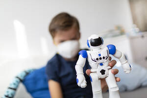 Junge mit Maske, der während der Corona-Krise mit seinem neuen, einzigen Freund, einem Roboter, in seinem Zimmer spielt - HMEF00825