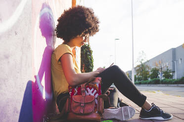 Junge Frau mit Afrofrisur sitzt an einer Graffiti-Wand und benutzt ein Smartphone - MEUF00219