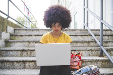 Junge Frau mit Afrofrisur benutzt Laptop auf einer Treppe in der Stadt - MEUF00228