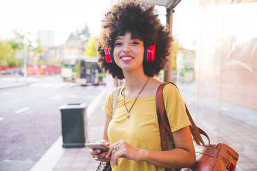 Glückliche junge Frau mit Afrofrisur, die mit Kopfhörern in der Stadt Musik hört - MEUF00231