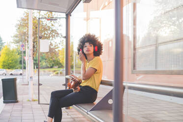 Junge Frau mit Afrofrisur, die an einer Bushaltestelle in der Stadt mit Kopfhörern Musik hört - MEUF00232