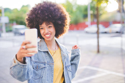 Glückliche junge Frau mit Afrofrisur macht ein Selfie in der Stadt - MEUF00238