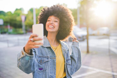 Glückliche junge Frau mit Afrofrisur macht ein Selfie in der Stadt - MEUF00239