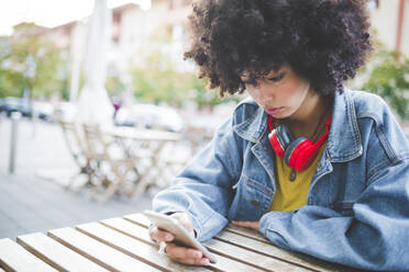 Junge Frau mit Afrofrisur benutzt ihr Smartphone in einem Café im Freien in der Stadt - MEUF00245