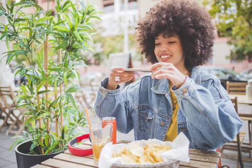 Smiling young woman with afro hairdo taking smartphone picture at an outdoor cafe in the city - MEUF00248