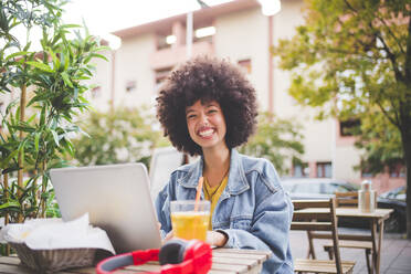 Happy young woman with afro hairdo using laptop at an outdoor cafe in the city - MEUF00252