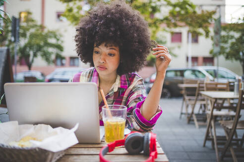 Junge Frau mit Afrofrisur, die in einem Straßencafé in der Stadt einen Laptop benutzt - MEUF00254