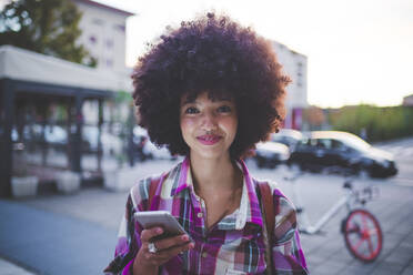 Porträt einer selbstbewussten jungen Frau mit Afrofrisur in der Stadt in der Abenddämmerung - MEUF00258