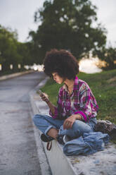 Junge Frau mit Afrofrisur sitzt auf einem Bordstein und benutzt ein Smartphone - MEUF00261