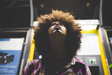Junge Frau mit Afrofrisur am Fahrkartenautomaten in der Nacht, die nach oben schaut - MEUF00273