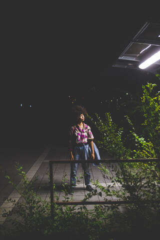 Junge Frau mit Afrofrisur steht nachts auf einem Bahnsteig, lizenzfreies Stockfoto