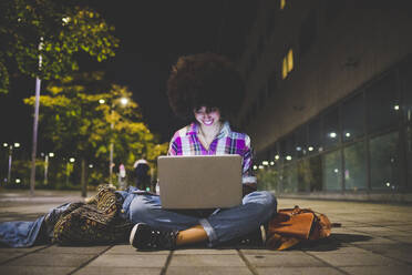 Junge Frau mit Afrofrisur sitzt auf dem Boden und benutzt einen Laptop in der Stadt bei Nacht - MEUF00275