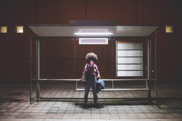 Young woman with afro hairdo using smartphone at bus stop in the city - MEUF00277