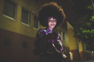 Junge Frau mit Afrofrisur, die nachts in der Stadt ihr Smartphone benutzt - MEUF00279