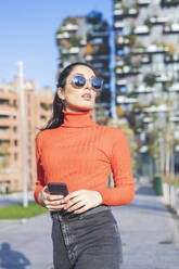 Schöne Frau trägt Sonnenbrille und hält Smartphone in der Stadt - MEUF00284