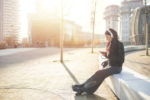 Schöne Frau, die auf einer Bank sitzt und ihr Smartphone benutzt, um Musik zu hören, lizenzfreies Stockfoto