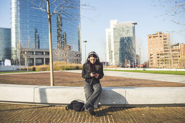 Schöne Frau, die auf einer Bank sitzt und ihr Smartphone benutzt, um Musik zu hören - MEUF00298
