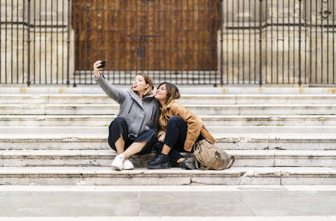 Zwei glückliche junge Frauen sitzen auf einer Treppe in der Stadt und machen ein Selfie, lizenzfreies Stockfoto