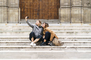 Zwei glückliche junge Frauen sitzen auf einer Treppe in der Stadt und machen ein Selfie - DGOF00571