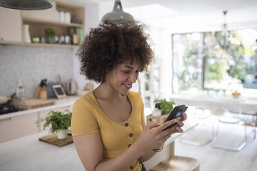 Smiling young woman using smart phone in kitchen - HOXF06063