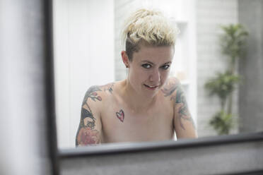 Portrait confident nude woman with tattooed shoulders at bathroom mirror - HOXF06124