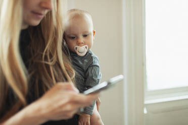 Neugieriges Baby-Mädchen mit Schnuller, das seine Mutter bei der Benutzung eines Smartphones beobachtet - HOXF06143