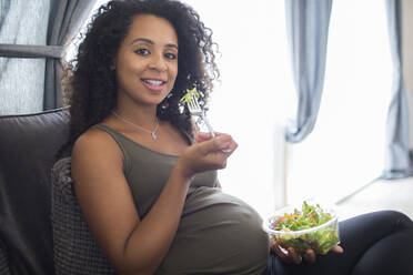 Portrait lächelnde junge schwangere Frau isst Salat auf Sofa - HOXF06150