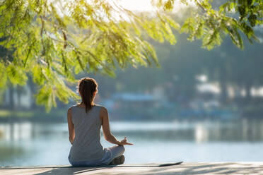 Rear View Of Young Woman Meditating While Sitting By Lake At Park - EYF01730