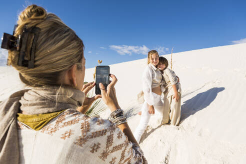 Eine Frau fotografiert ihre Kinder mit einem Smartphone in einer weißen Sanddünenlandschaft unter blauem Himmel. - MINF14323