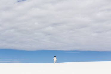 Ein kleiner Junge, der durch weiße Sanddünen spaziert, ein Streifen blauer Himmel am Horizont und dicke Wolken. - MINF14332