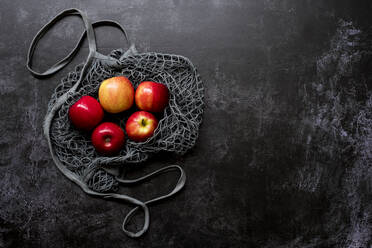 High angle close up of red apples in grey net bag on black background. - MINF14458